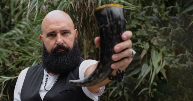 A bearded man making a toast gesture with a drinking horn on hand.