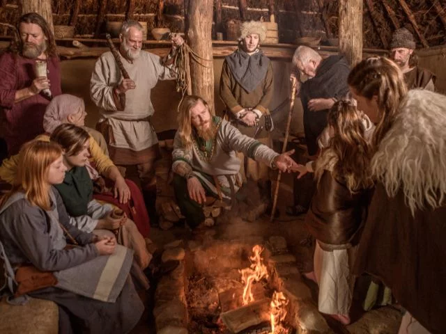 A group of vikings gathered and huddled in house with a fireplace at the center.