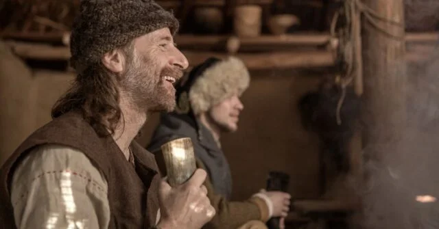 A viking man holding and drinking from a horn in a gathering.