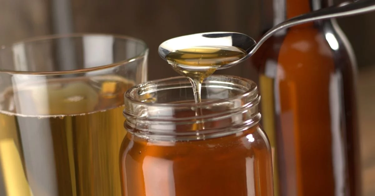 A glass with mead and a jar of honey with a spoon in it.